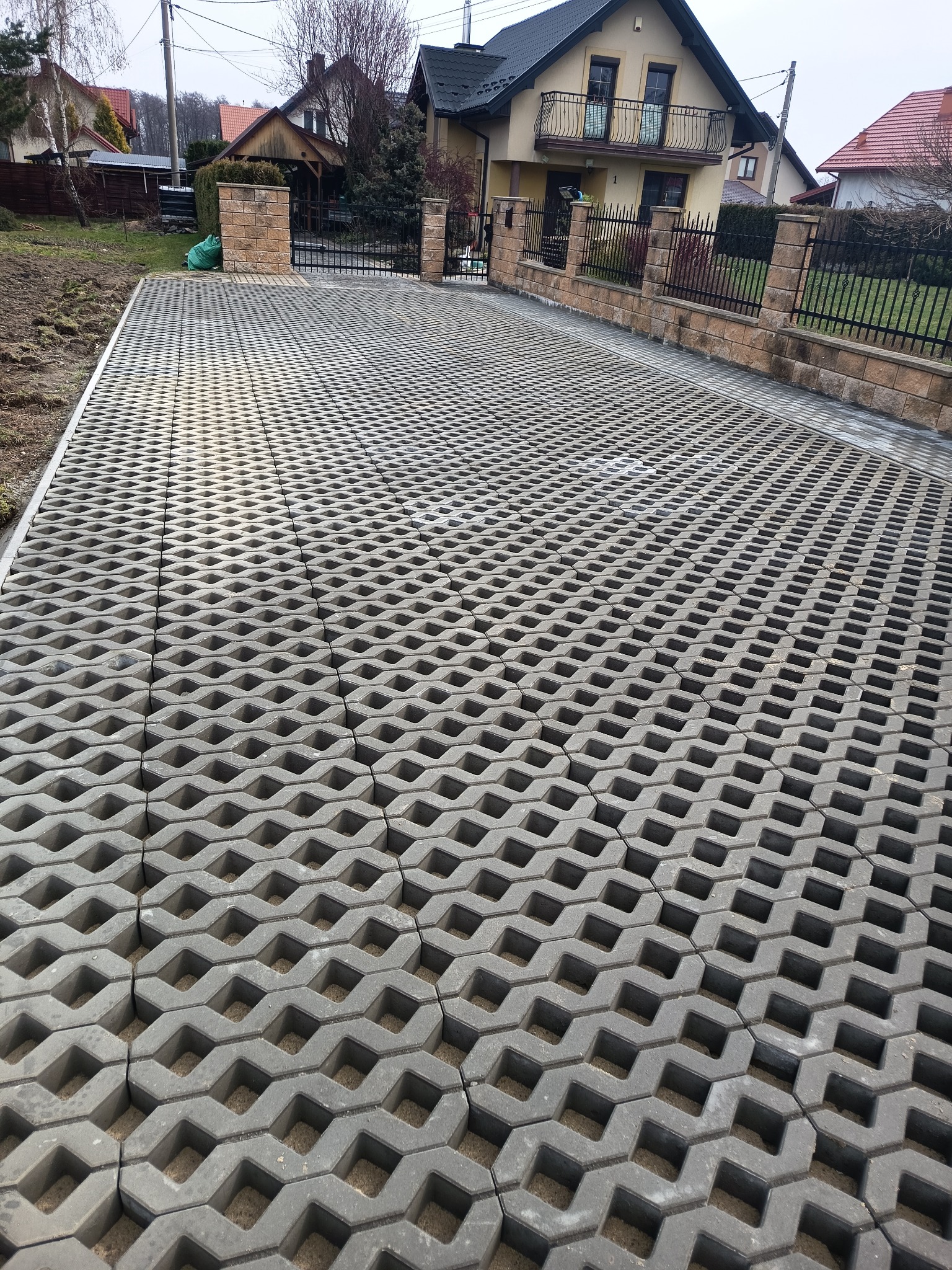 Szara, ażurowa kostka na podjeździe, widok z góry. W tle dom z balkonem i ogrodzenie z kamiennymi słupkami. Realizacja na posesji.