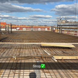 ROS-BUD - Zbrojenie stropu na budowie z widokiem na miasto w tle. Siatka z prętów stalowych ułożona na deskowaniu. Widoczne niebo z chmurami.