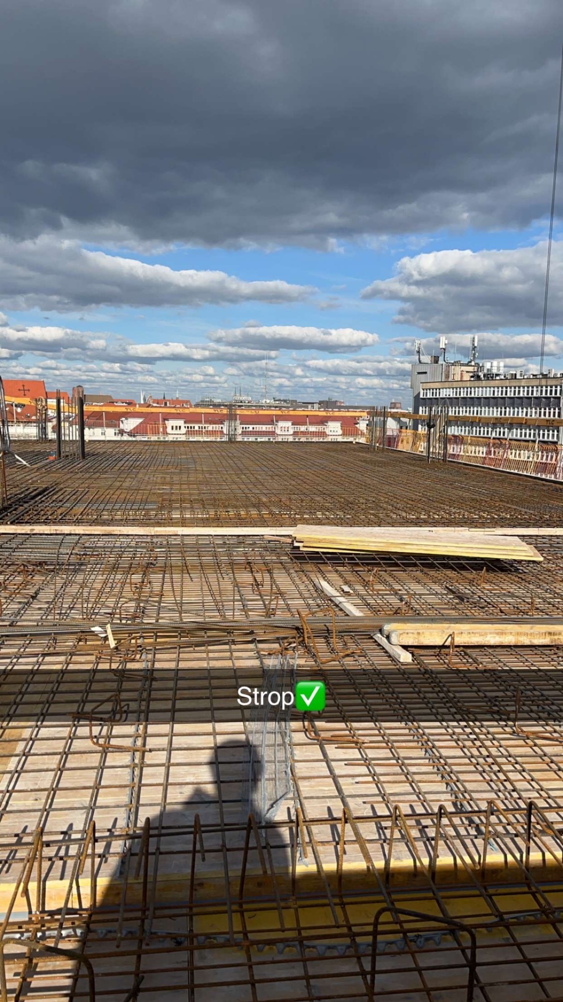 Zbrojenie stropu na budowie z widokiem na miasto w tle. Siatka z prętów stalowych ułożona na deskowaniu. Widoczne niebo z chmurami.