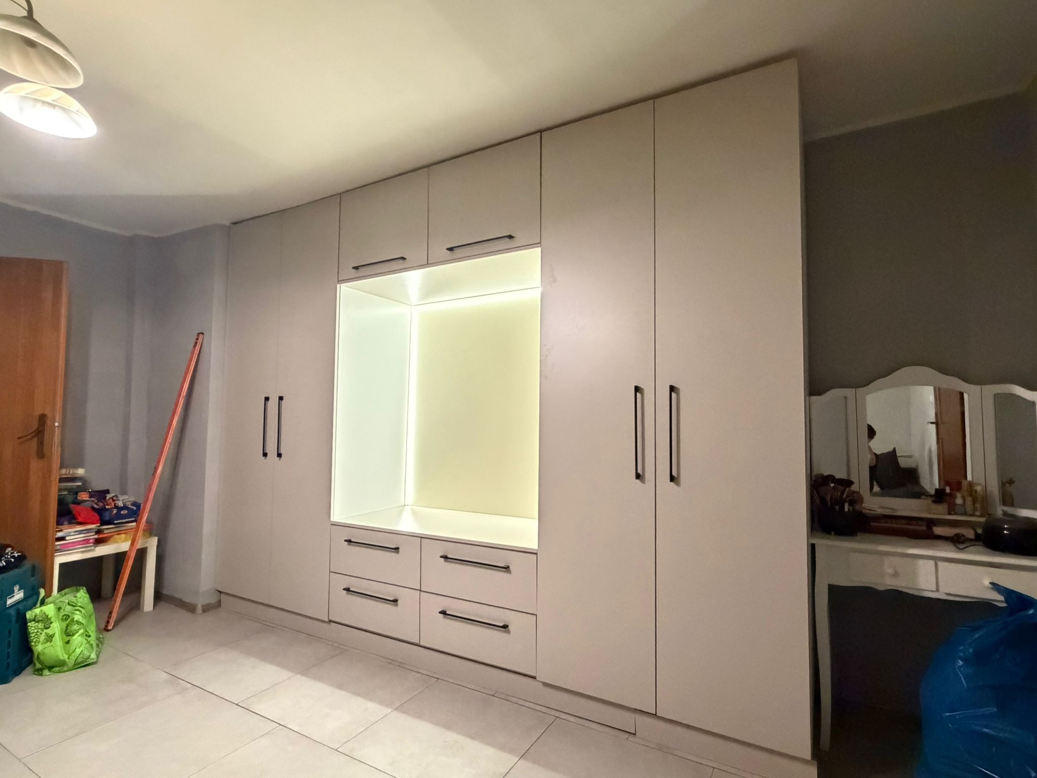 Biała szafa na wymiar z oświetleniem LED wnęki, czarne uchwyty, jasne płytki podłogowe, toaletka z lustrem w tle. Realizacja szafy wnękowej.