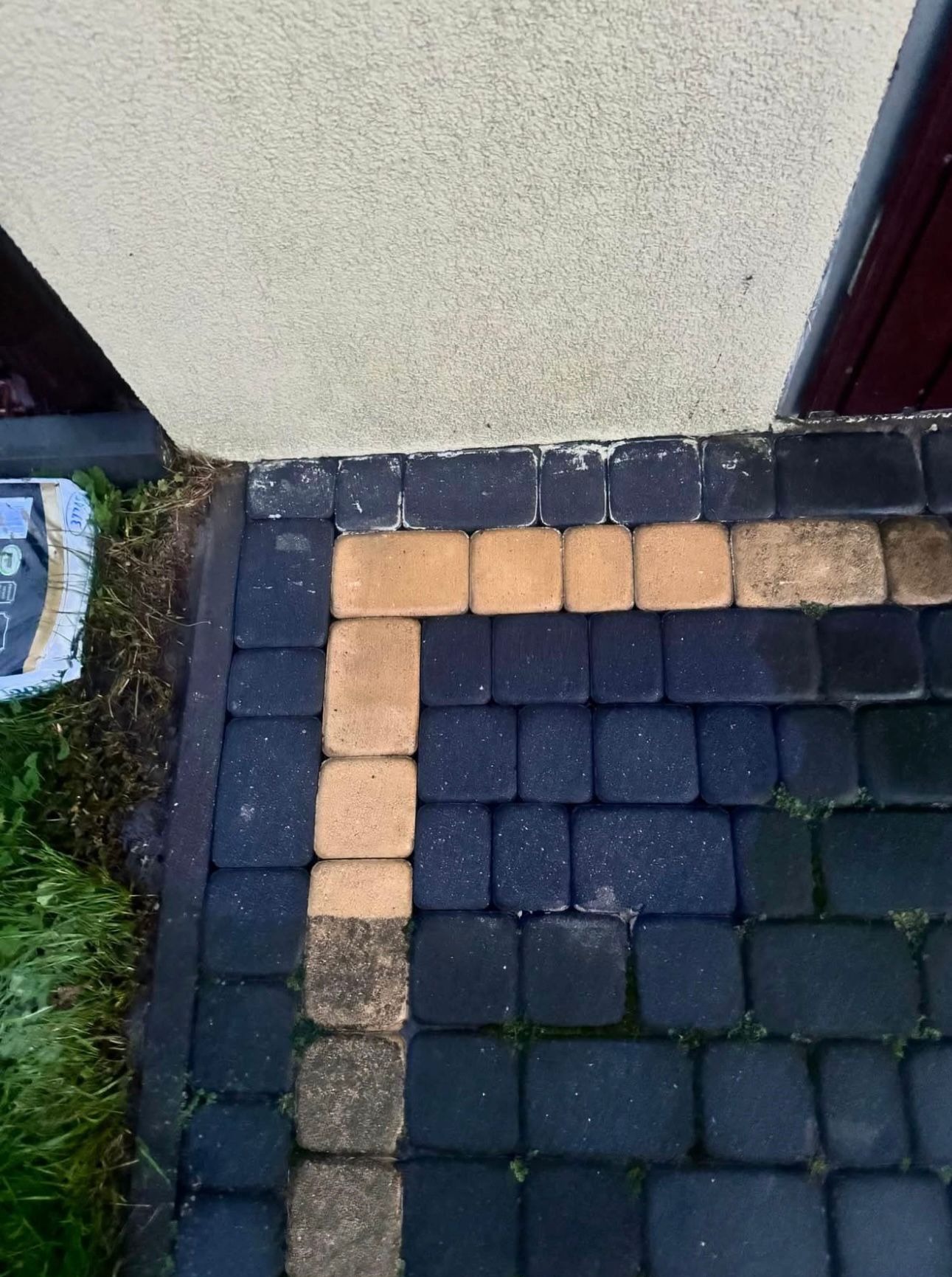 Ułożona kostka brukowa w dwóch kolorach (czarny i beżowy) przy ścianie budynku. Fragment kostki jest jeszcze zabrudzony, co sugeruje etap czyszczenia.