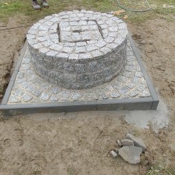 MATBRUK - Studnia z kostki granitowej, okrągła konstrukcja na kwadratowej podstawie, otoczona ziemią i trawą. Widoczny błękitny kubeł i zwinięty kabel.