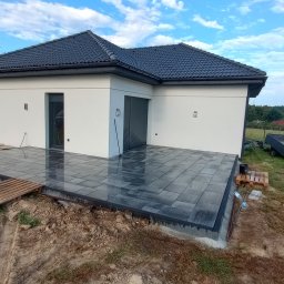 MATBRUK - Nowoczesny taras z ciemnej kostki brukowej przy białym domu z szarą stolarką. Widoczne narzędzia i materiały budowlane. Tło stanowi zieleń ogrodu i błękitne niebo.