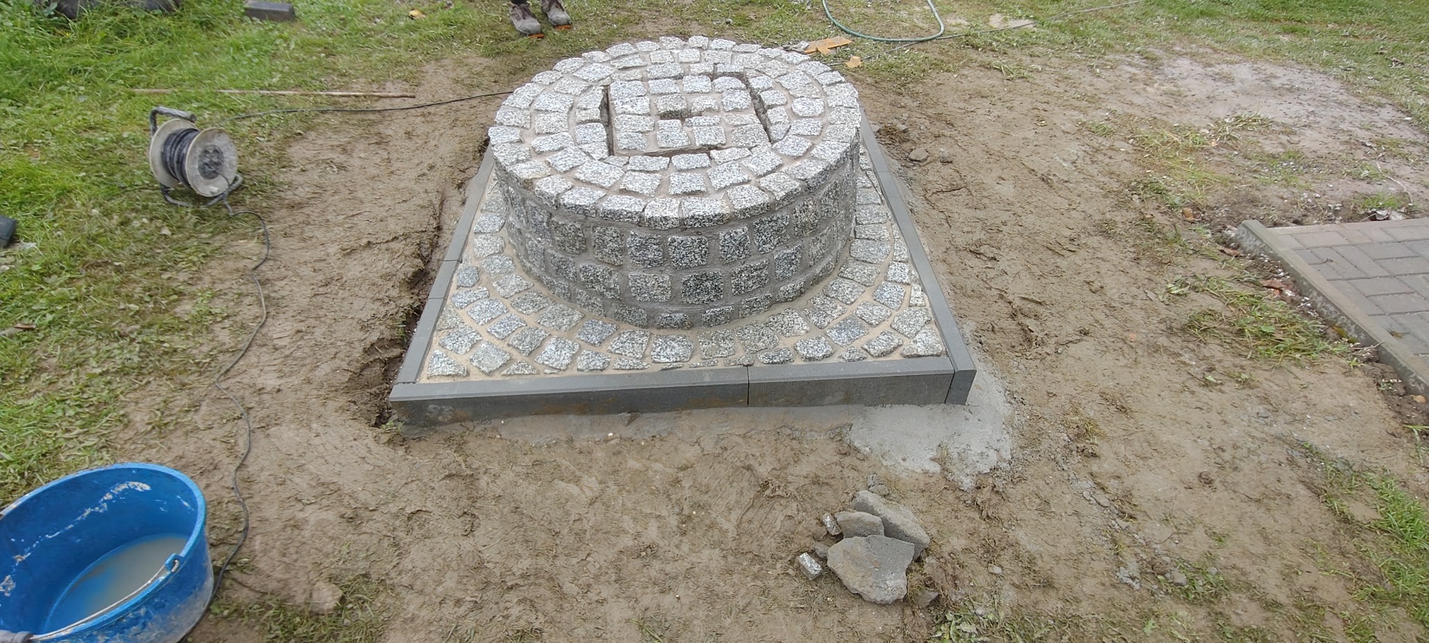 Studnia z kostki granitowej, okrągła konstrukcja na kwadratowej podstawie, otoczona ziemią i trawą. Widoczny błękitny kubeł i zwinięty kabel.