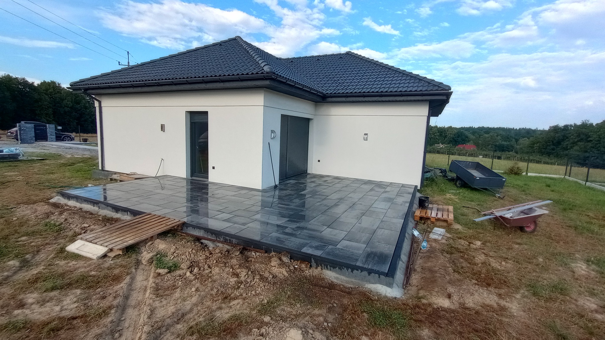 Nowoczesny taras z ciemnej kostki brukowej przy białym domu z szarą stolarką. Widoczne narzędzia i materiały budowlane. Tło stanowi zieleń ogrodu i błękitne niebo.