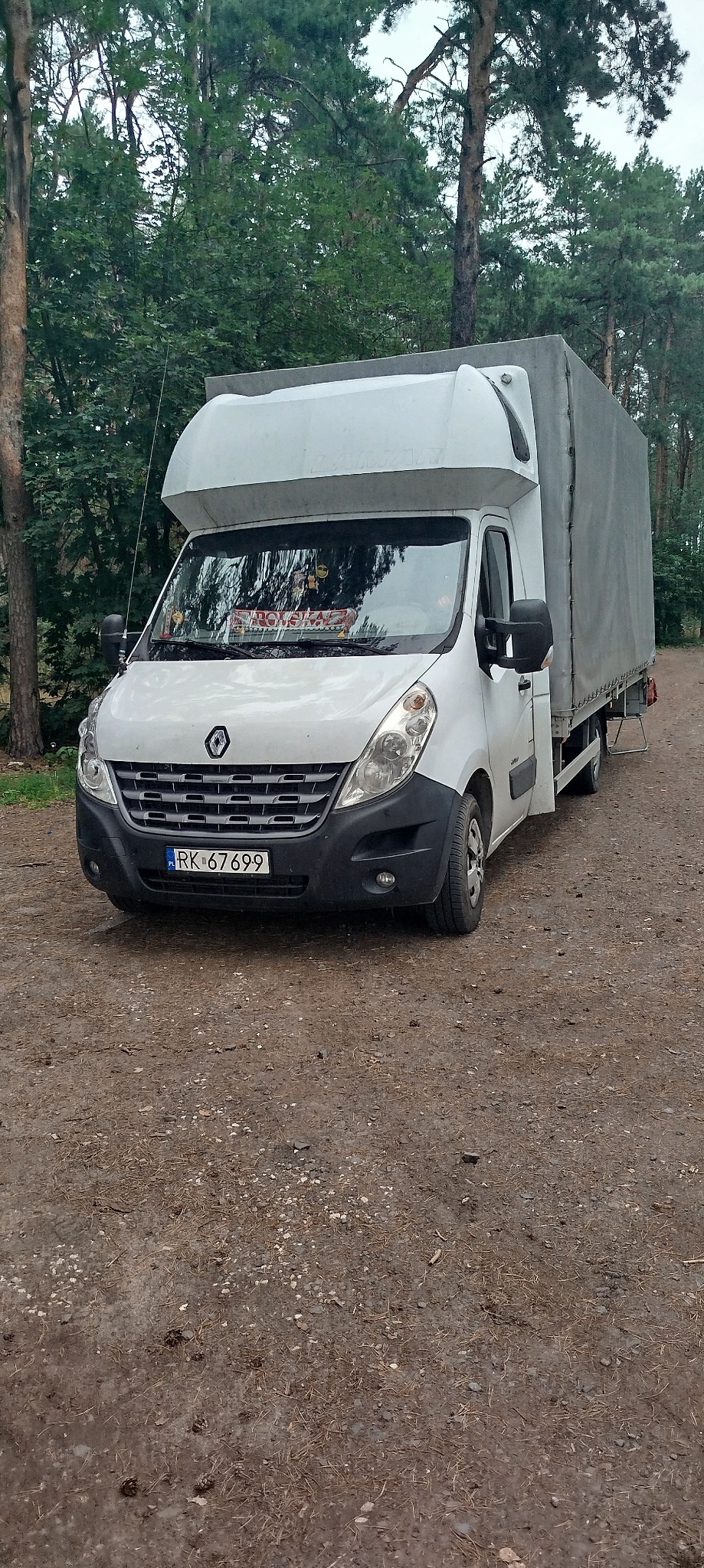 Biały Renault Master z plandeką na leśnej drodze. Widoczna tablica rejestracyjna i naklejka 'Polska' na przedniej szybie. Ujęcie z dołu, drzewa w tle.