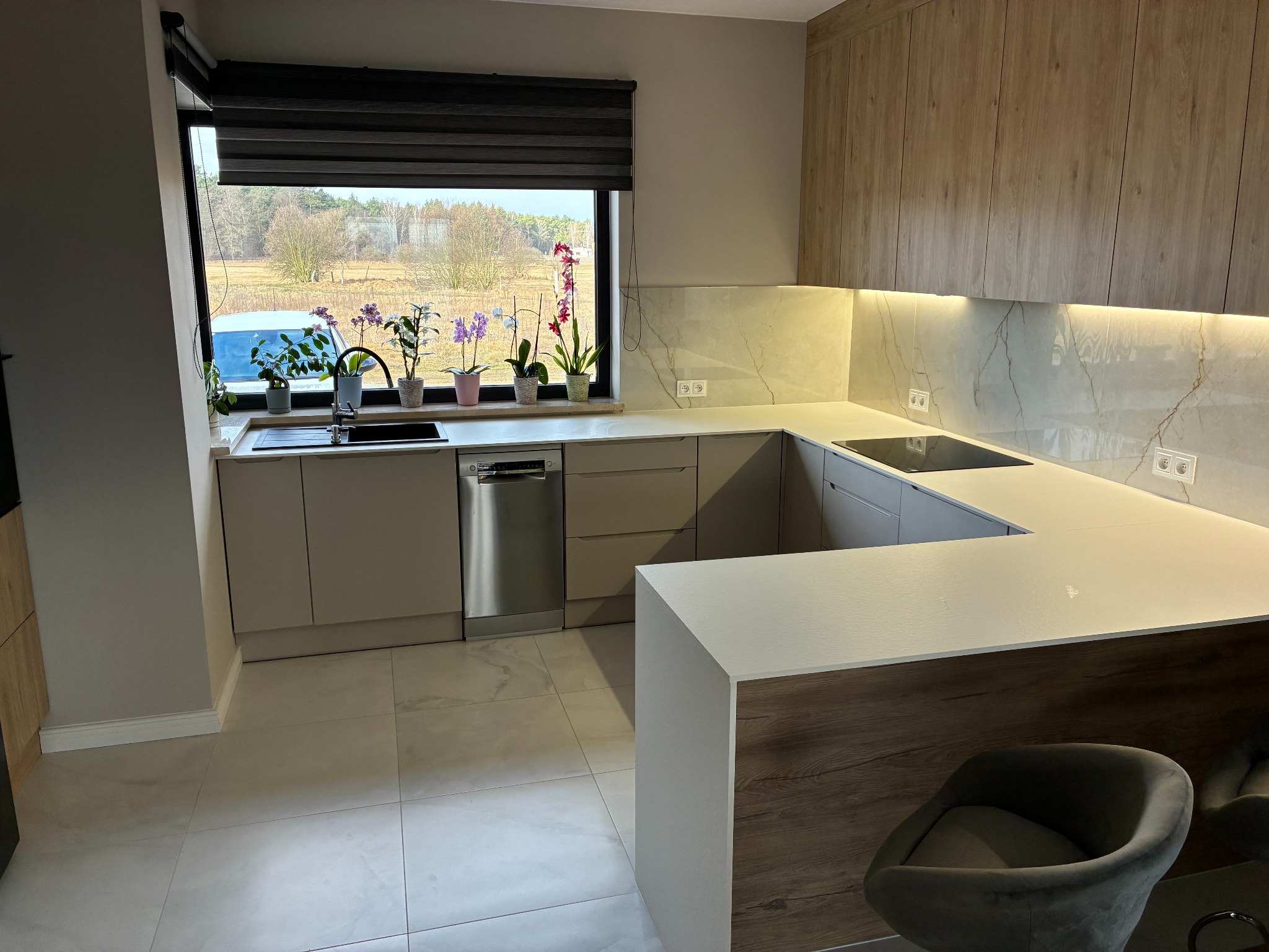 Nowoczesna kuchnia w kształcie litery U z wyspą, beżowe fronty, drewniane akcenty, marmurowy backsplash, oświetlenie LED i kwiaty w oknie z widokiem na zewnątrz.
