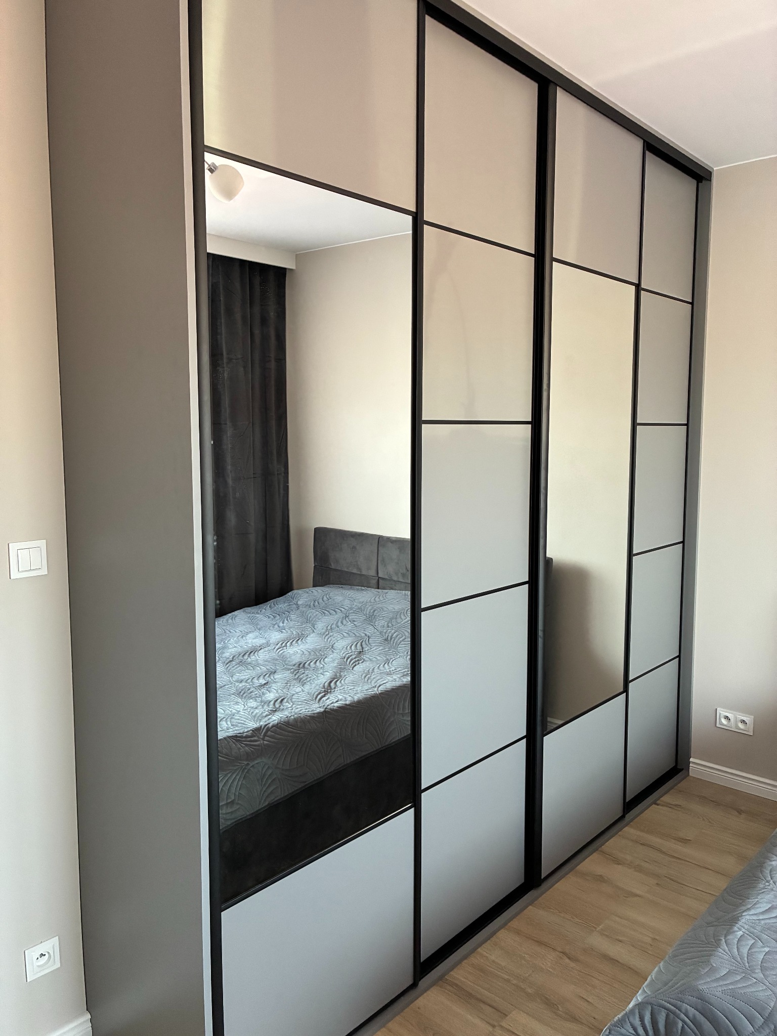 Nowoczesna szafa wnękowa z przesuwnymi drzwiami, dzielonymi na kwadratowe panele w odcieniach szarości i z lustrem, odbijającym wnętrze sypialni z łóżkiem.