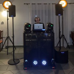 Music Jedi DJ - DJ z laptopem za konsoletą, otoczony głośnikami i reflektorami. Scena oświetlona ciepłym światłem, tworząca klimat imprezy. Widoczne elementy nagłośnienia.