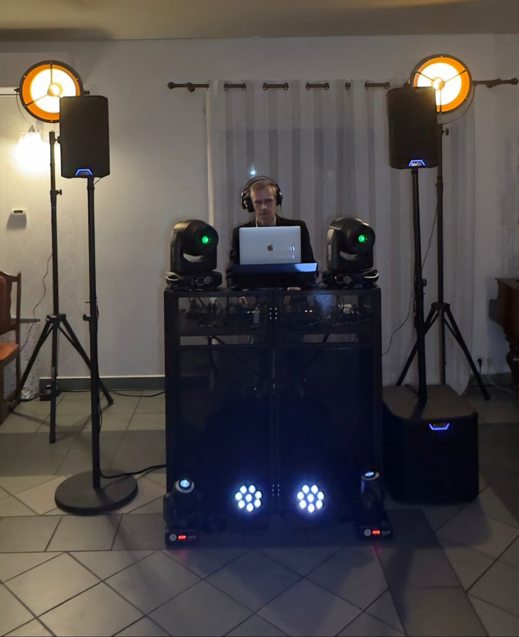 DJ z laptopem za konsoletą, otoczony głośnikami i reflektorami. Scena oświetlona ciepłym światłem, tworząca klimat imprezy. Widoczne elementy nagłośnienia.