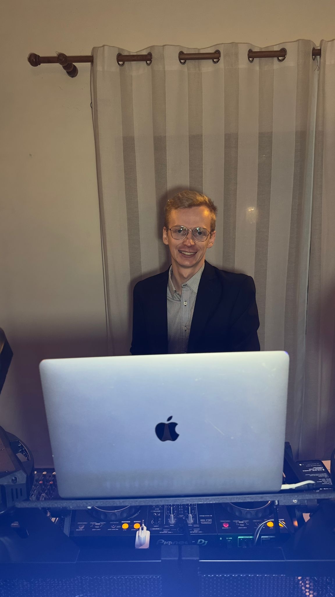Uśmiechnięty DJ w okularach, w marynarce, za konsoletą Pioneer DJ i laptopem Apple, na tle zasłon. Gotowy do występu na imprezie.