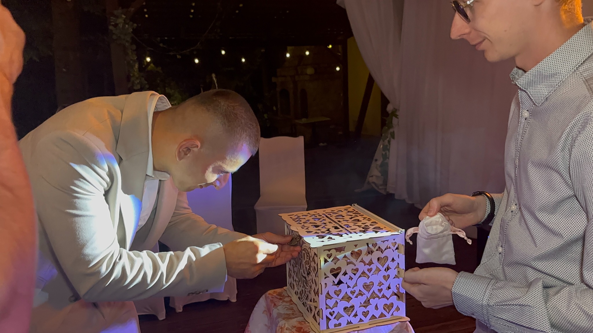 Moment wrzucania prezentu do ozdobnej, ażurowej skrzynki na weselu. Mężczyzna w garniturze wkłada kopertę, drugi trzyma woreczek z upominkiem. Dekoracje i oświetlenie w tle.