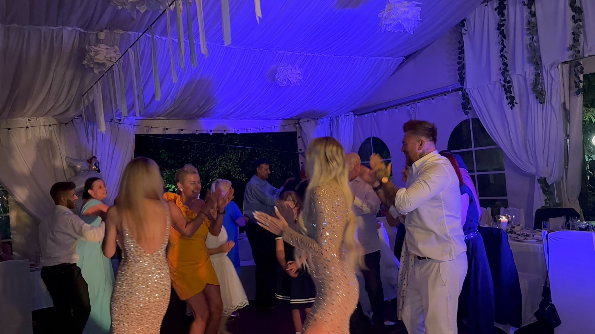 Rozbawieni goście weselni tańczą w udekorowanym namiocie, oświetlonym niebieskim światłem. W tle widoczne stoły i krzesła, a także dekoracje sufitowe.