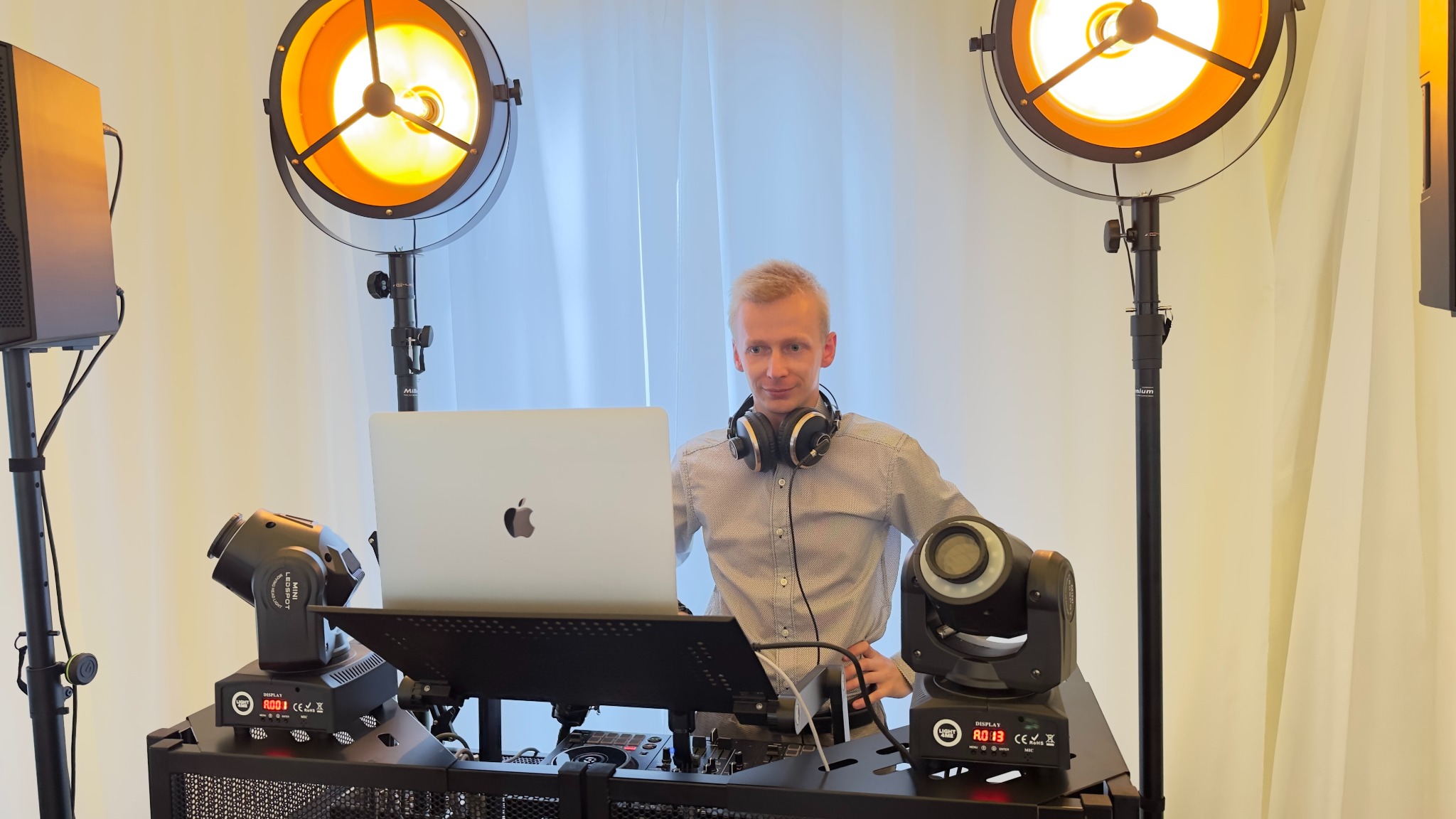 DJ z laptopem i słuchawkami, otoczony sprzętem nagłośnieniowym i oświetleniowym, w tle jasna zasłona. Widoczne reflektory i głośniki.