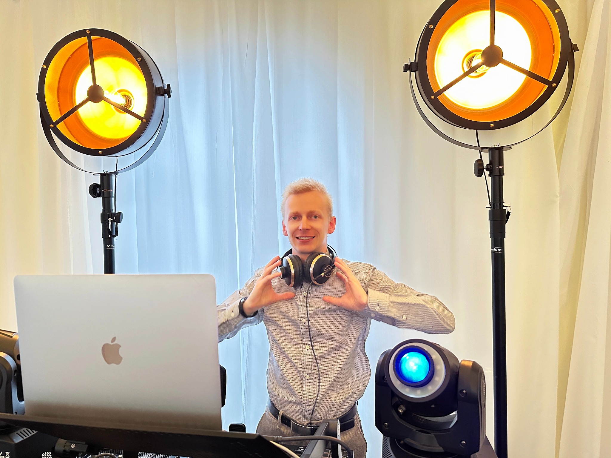 Uśmiechnięty DJ z słuchawkami na szyi, robiący serce dłońmi, otoczony sprzętem: laptop, reflektory i ruchoma głowa. Białe zasłony w tle.