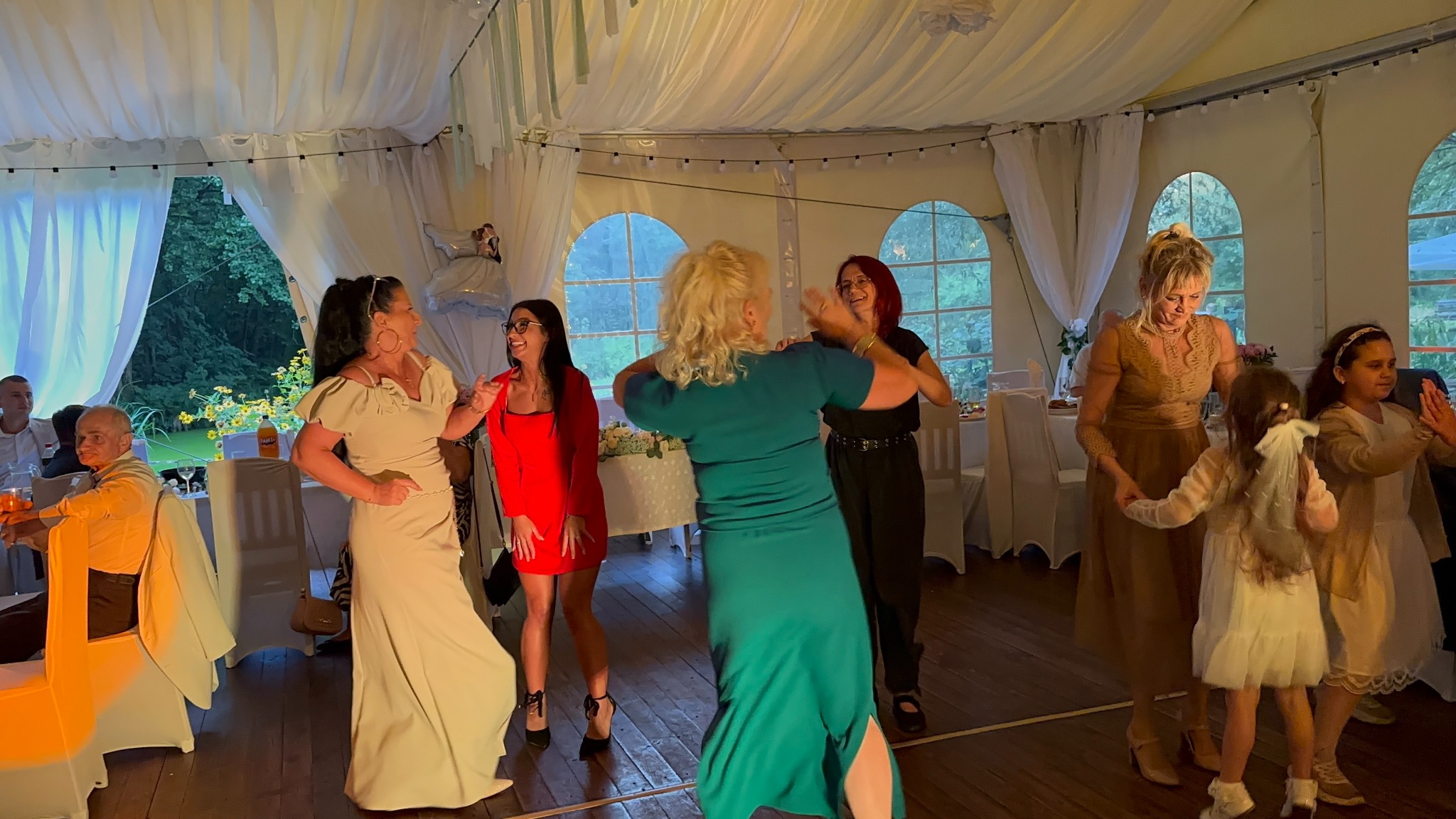 Roztańczone kobiety w eleganckim namiocie podczas przyjęcia. Dekoracje kwiatowe, białe zasłony i okna z widokiem na zieleń tworzą radosną atmosferę.