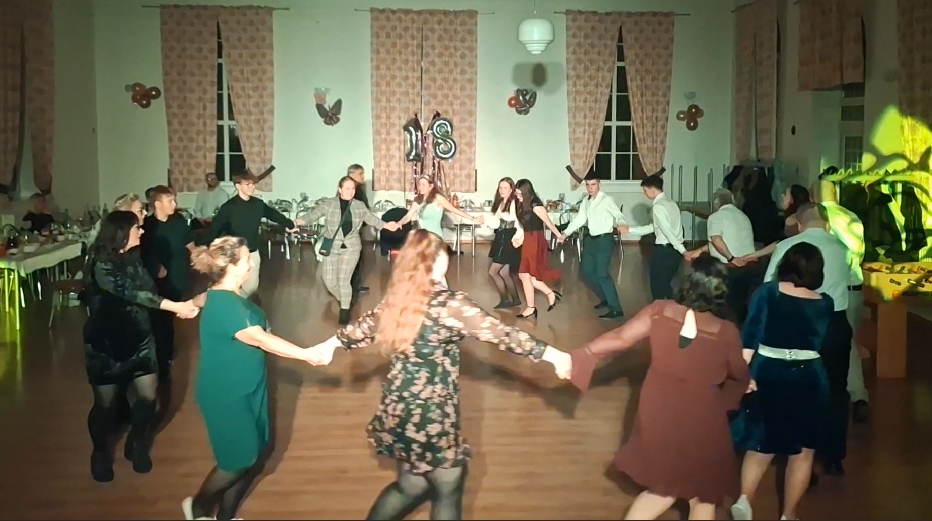 Uczestnicy osiemnastki tańczą w kręgu na parkiecie sali balowej, w tle dekoracje urodzinowe i nakryte stoły. Żółte oświetlenie sceniczne.