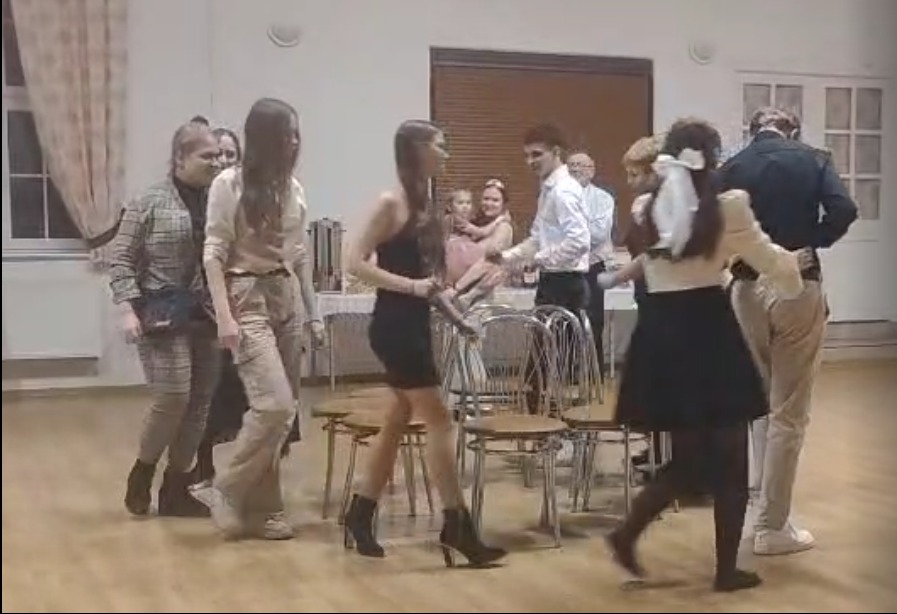 Zabawa taneczna w sali: grupa osób tańczy wokół krzeseł, w tle stół z przekąskami i obserwujący. Uczestnicy w różnorodnych strojach, dynamiczne ujęcie.