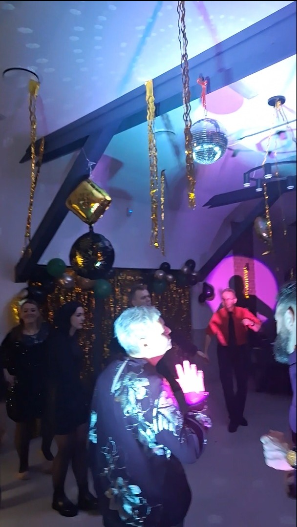 Impreza w sali z dekoracjami: balony, kule dyskotekowe, girlandy. W tle tańczące osoby, oświetlenie sceniczne w odcieniach fioletu i niebieskiego.