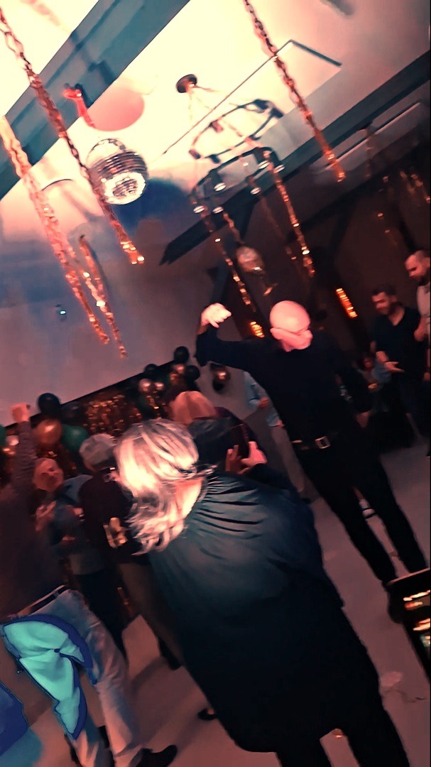 Roztańczony tłum na imprezie z dekoracjami: balony, łańcuchy i kula dyskotekowa. Mężczyzna w czerni z uniesioną ręką w centrum uwagi, dynamiczne ujęcie.