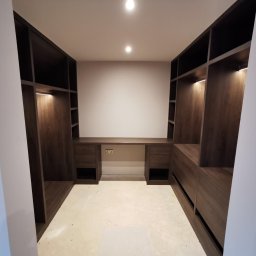 Grzegorz Augustynek - Funkcjonalna garderoba z biurkiem w zabudowie, wykonana z ciemnego drewna. Oświetlenie LED podkreśla nowoczesny design. Widok z perspektywy wejścia.