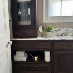 Grzegorz Augustynek - Ciemna, drewniana szafka łazienkowa z marmurowym blatem, lustrem i rośliną. Widoczne ręczniki, złoty dzbanek i mozaika na podłodze. Okno z mleczną szybą w tle.