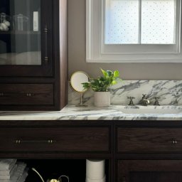 Grzegorz Augustynek - Ciemna, drewniana szafka łazienkowa z marmurowym blatem, złotą konewką i lustrem na tle okna z mlecznymi szybami. Elegancki detal wyposażenia wnętrza.