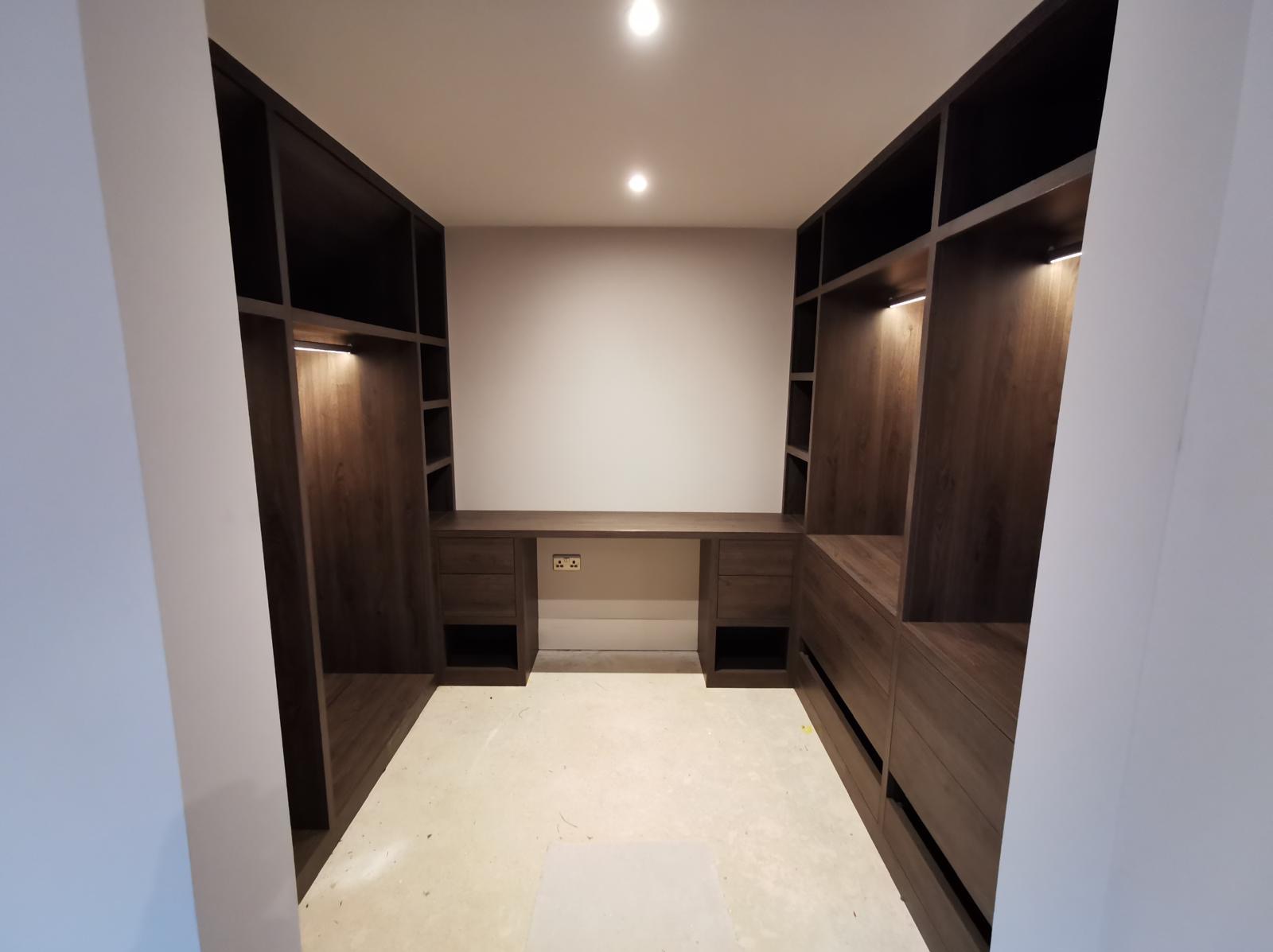 Funkcjonalna garderoba z biurkiem w zabudowie, wykonana z ciemnego drewna. Oświetlenie LED podkreśla nowoczesny design. Widok z perspektywy wejścia.