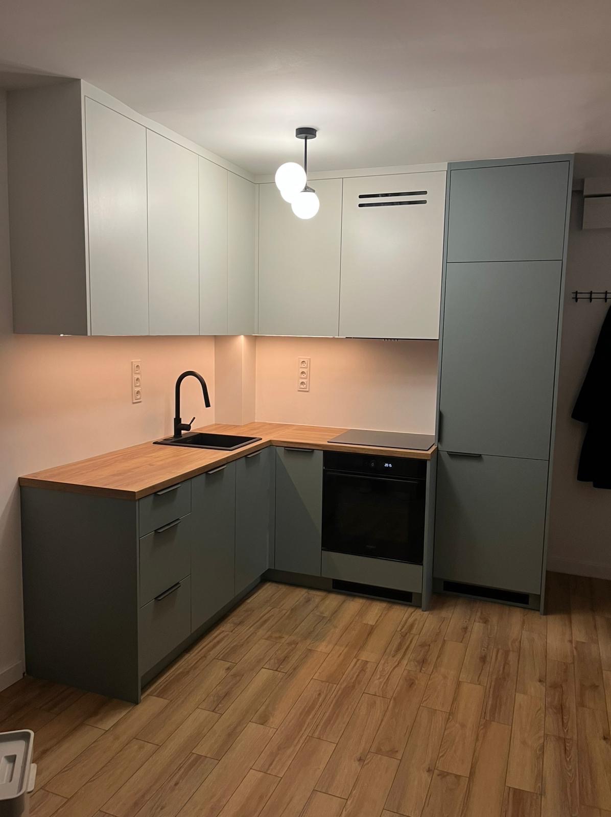 Szara kuchnia na wymiar z drewnianym blatem i czarną armaturą. Górne szafki białe, oświetlenie LED pod szafkami. Na podłodze panele imitujące drewno.