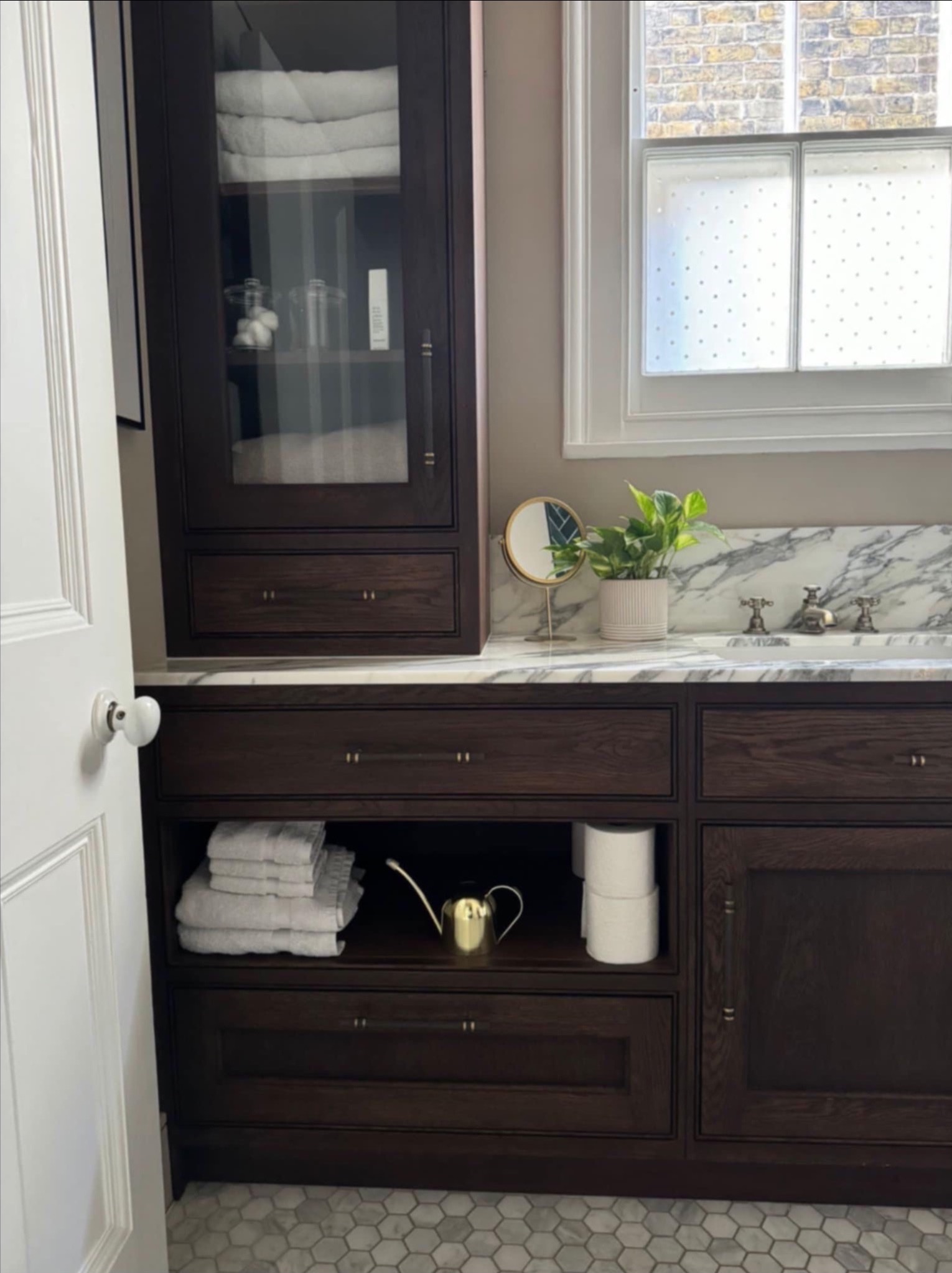 Ciemna, drewniana szafka łazienkowa z marmurowym blatem, lustrem i rośliną. Widoczne ręczniki, złoty dzbanek i mozaika na podłodze. Okno z mleczną szybą w tle.