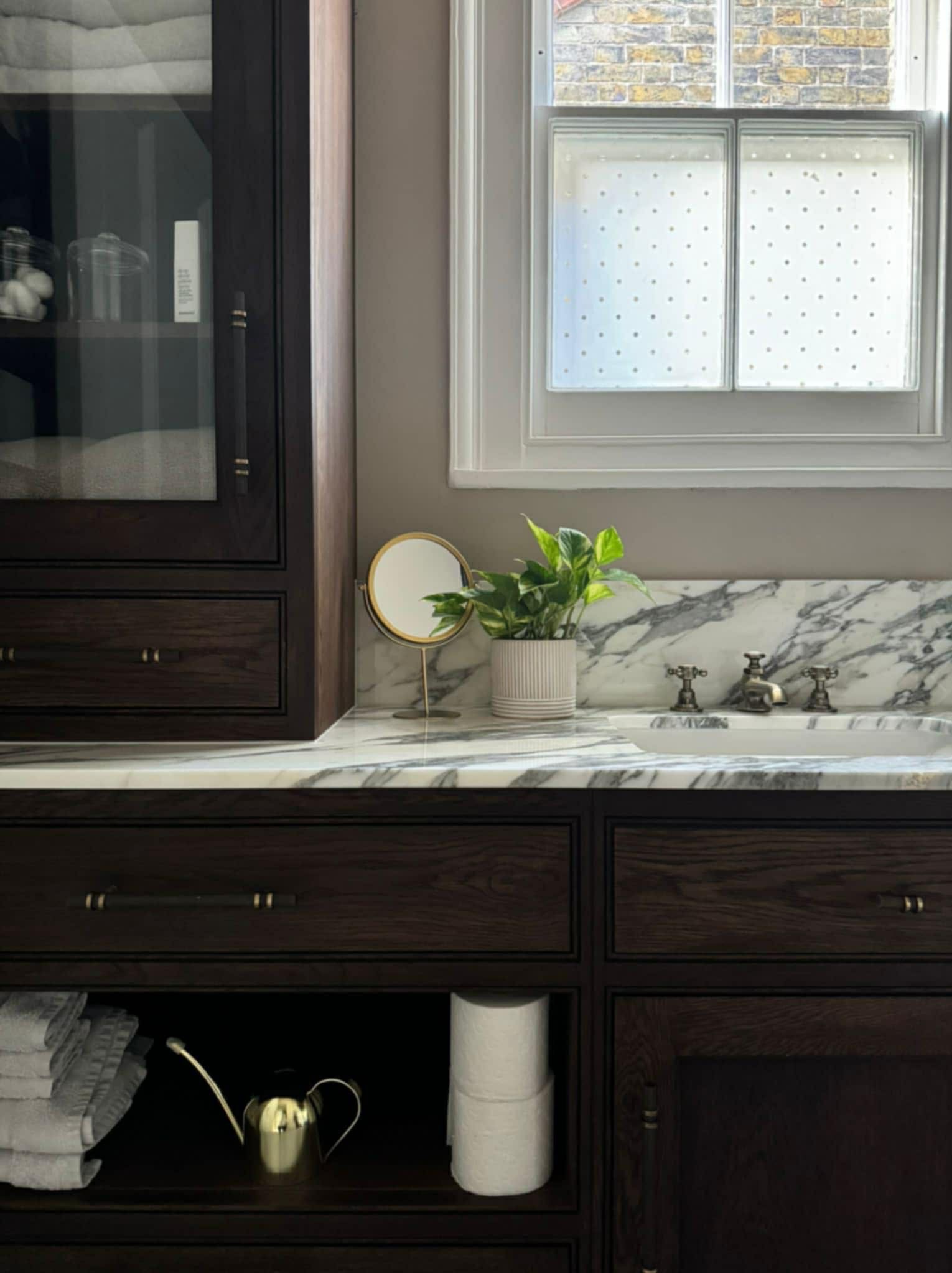 Ciemna, drewniana szafka łazienkowa z marmurowym blatem, złotą konewką i lustrem na tle okna z mlecznymi szybami. Elegancki detal wyposażenia wnętrza.