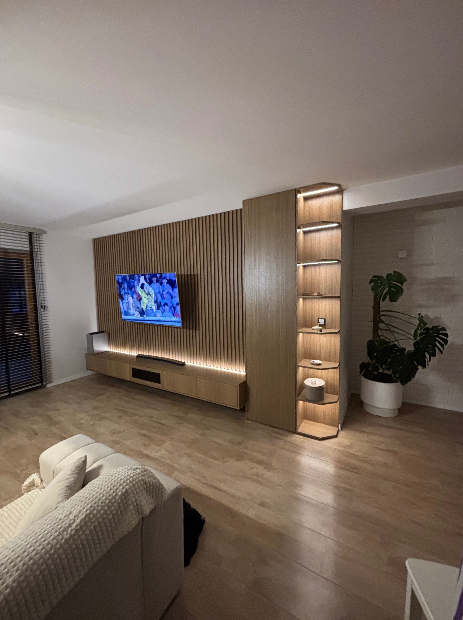 Nowoczesny salon z drewnianą zabudową TV z lamelami i podświetleniem LED, regał z półkami i rośliną doniczkową. Minimalistyczny design, ciepłe oświetlenie.
