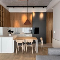 AWPLUS Architektura wnętrz Maja Staniszewska - Nowoczesna kuchnia z drewnianymi szafkami i szarymi akcentami, oświetlona wiszącą lampą w kształcie stożka. Stół z krzesłami na pierwszym planie, widok z salonu.