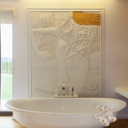 AWPLUS Architektura wnętrz Maja Staniszewska - Biała, wolnostojąca wanna z kwiatami orchidei na drewnianej podłodze. W tle dekoracyjna płaskorzeźba nad wanną i okno z widokiem na zieleń.