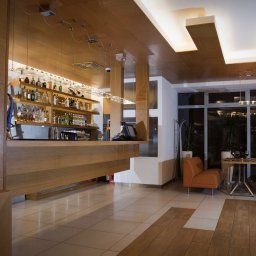 AWPLUS Architektura wnętrz Maja Staniszewska - Nowoczesne wnętrze baru z drewnianym wykończeniem, oświetleniem punktowym i butelkami alkoholu na półkach. Widoczne stoliki i krzesła dla gości.