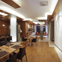 AWPLUS Architektura wnętrz Maja Staniszewska - Eleganckie wnętrze restauracji z drewnianymi stołami nakrytymi dla gości, oświetleniem sufitowym i dekoracyjną ścianą z logo.