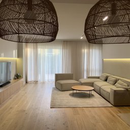 AWPLUS Architektura wnętrz Maja Staniszewska - Jasny salon z drewnianą podłogą, beżową sofą modułową, okrągłym stolikiem kawowym i dwoma dużymi, plecionymi lampami wiszącymi. Minimalistyczny design z akcentami naturalnymi.