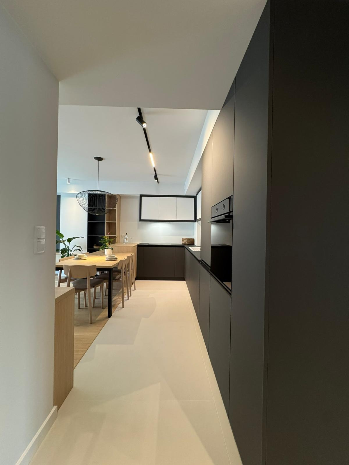 Nowoczesna kuchnia z jadalnią w minimalistycznym stylu. Ciemne szafki kuchenne kontrastują z jasną podłogą i stołem. Widoczne oświetlenie punktowe i designerska lampa.