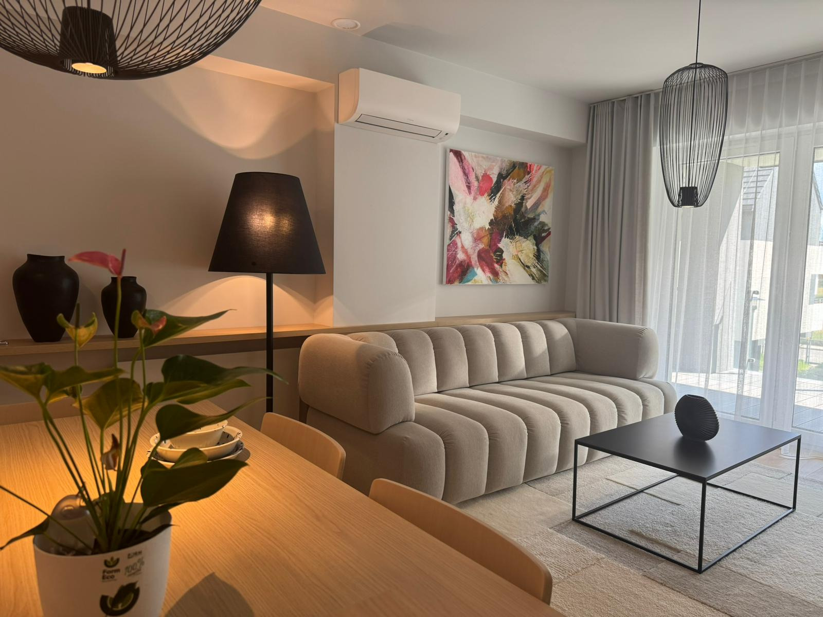 Jasne wnętrze: beżowa sofa z poduszkami, czarny stolik kawowy, abstrakcyjny obraz na ścianie, lampa z abażurem i roślina na pierwszym planie.