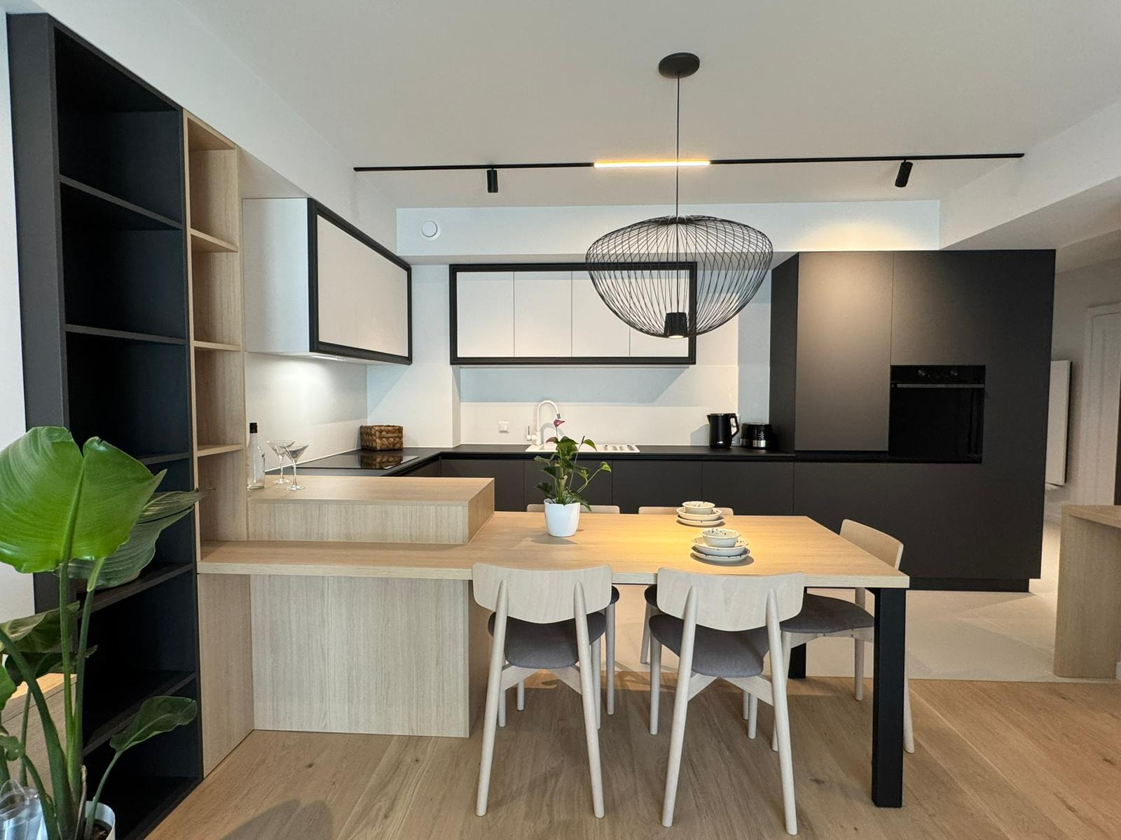 Nowoczesna kuchnia z jadalnią: czarne szafki, drewniany stół z krzesłami, designerska lampa wisząca. Minimalistyczny wystrój, roślina doniczkowa dodaje świeżości.