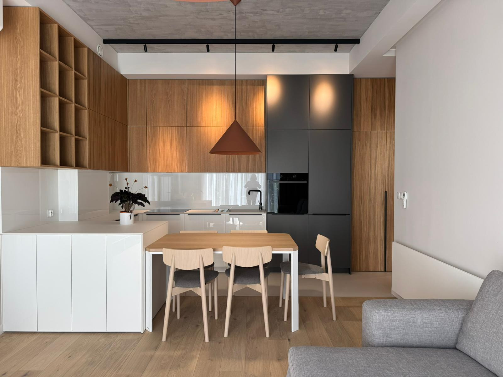 Nowoczesna kuchnia z drewnianymi szafkami i szarymi akcentami, oświetlona wiszącą lampą w kształcie stożka. Stół z krzesłami na pierwszym planie, widok z salonu.