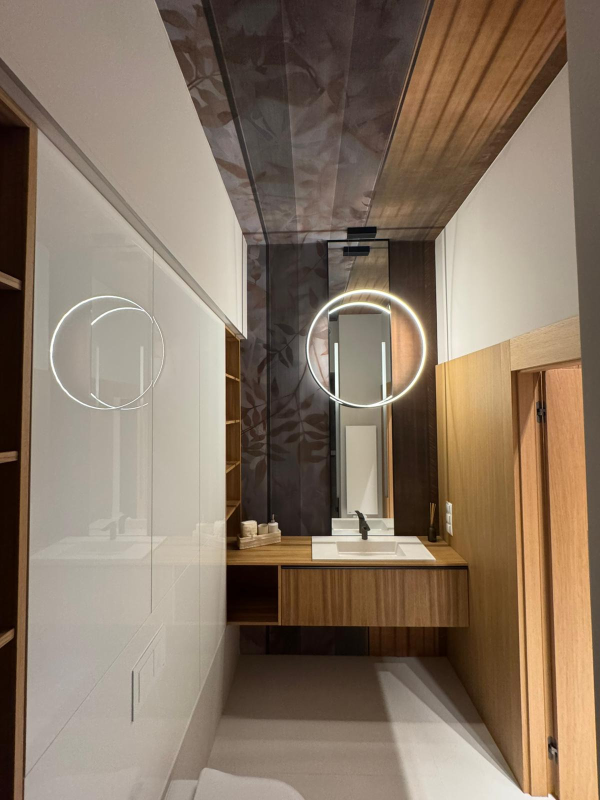 Nowoczesna łazienka z drewnianymi akcentami: szafka pod umywalką, lustro z podświetleniem LED i dekoracyjna tapeta na suficie z motywem liści.
