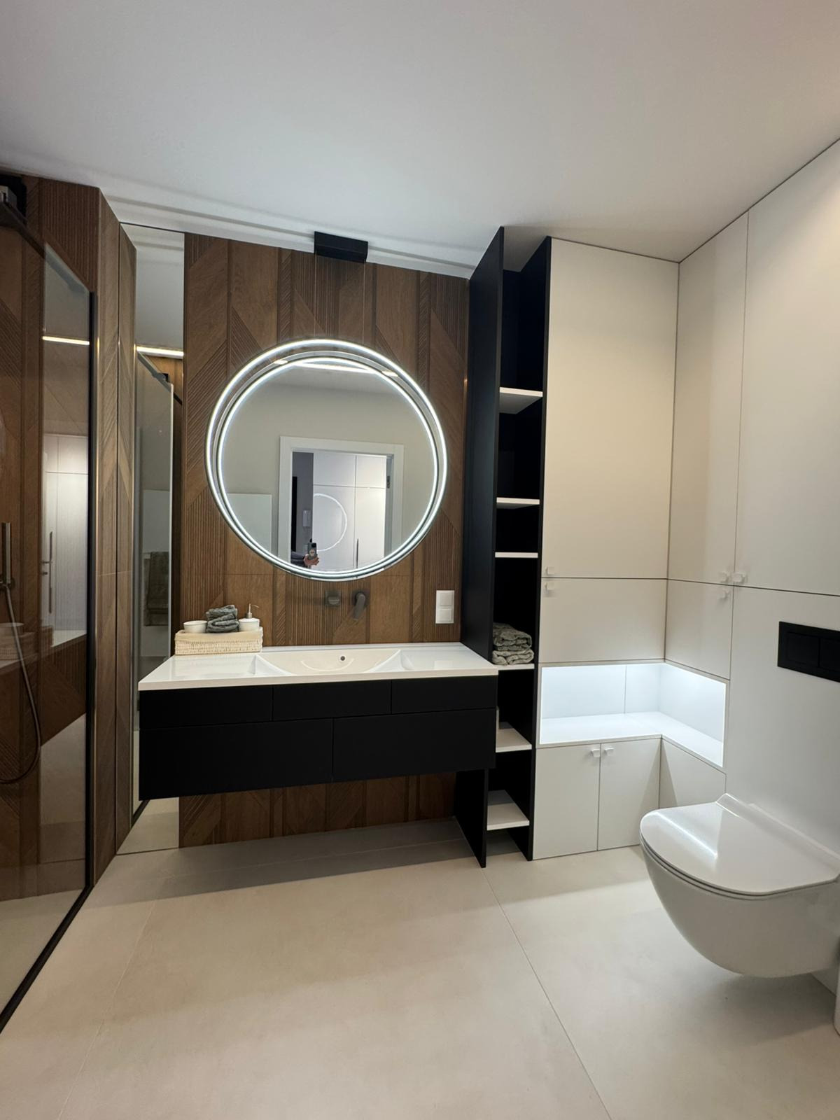 Nowoczesna łazienka z drewnianą ścianą, okrągłym lustrem z podświetleniem LED i czarną szafką pod umywalką. Minimalistyczny design z elementami bieli i czerni.