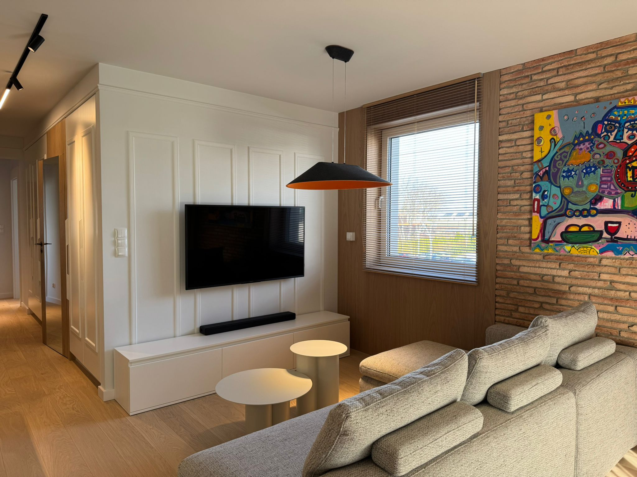 Jasne wnętrze salonu z ceglaną ścianą, nowoczesnym oświetleniem i minimalistycznymi meblami. Widoczny fragment sofy i stoliki kawowe.