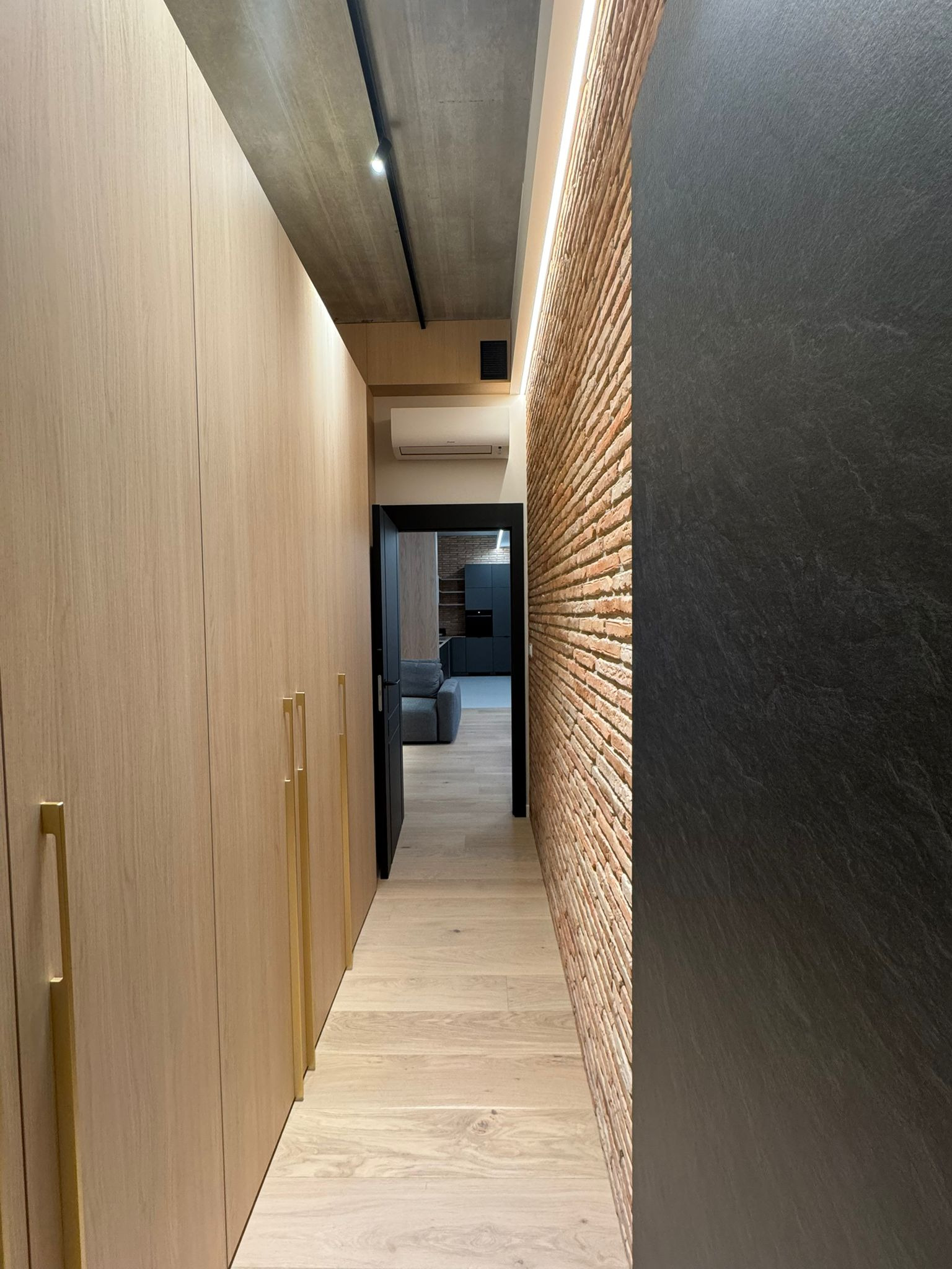 Wąski korytarz z zabudową szaf w jasnym drewnie i ścianą z cegły, oświetlony ledami. Widok na salon z szarą sofą. Minimalistyczny design.