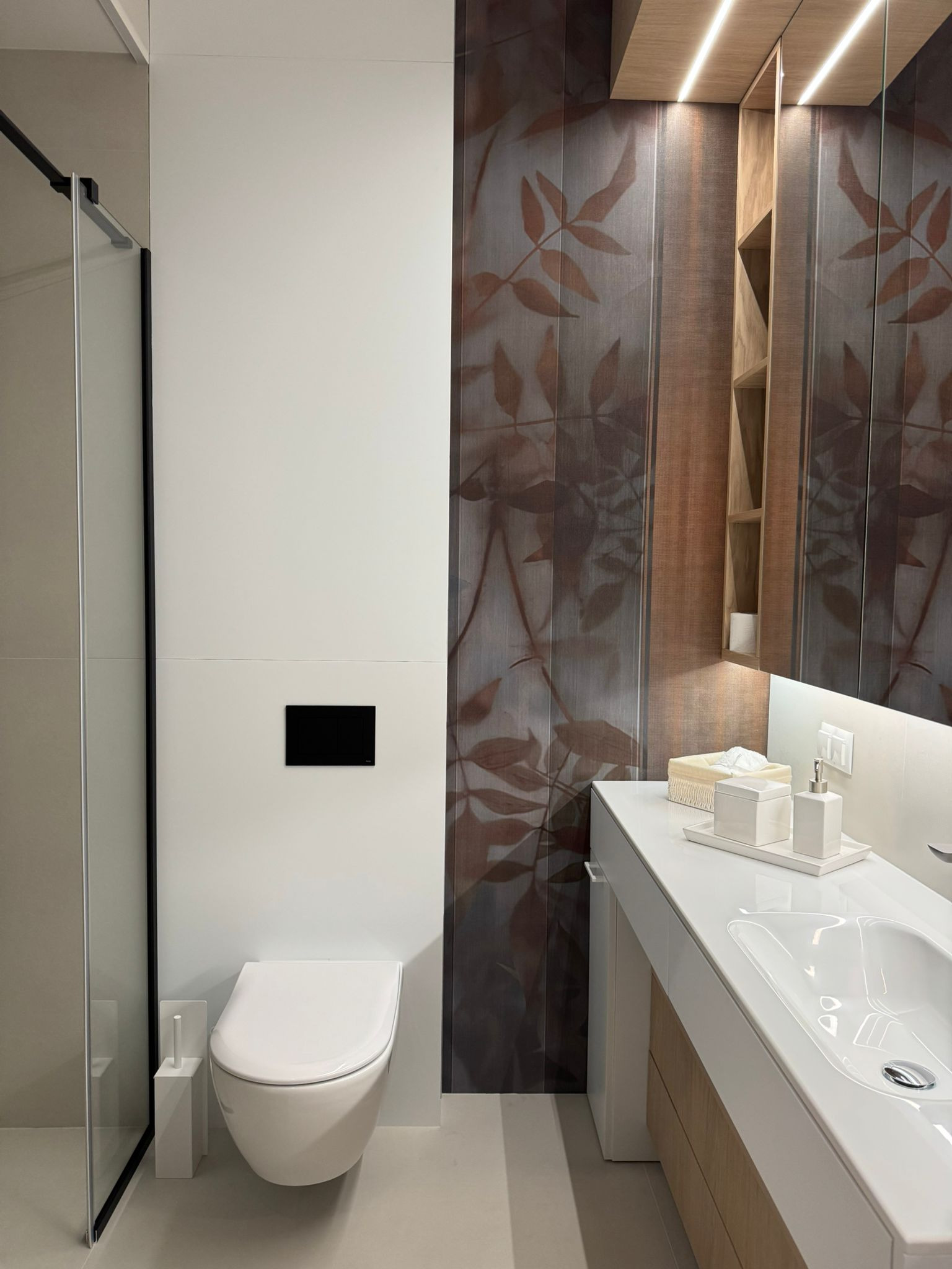 Nowoczesna łazienka z kabiną prysznicową, podwieszaną toaletą i umywalką wpuszczaną w blat. Dekoracyjna ściana z motywem liści i oświetlenie LED.