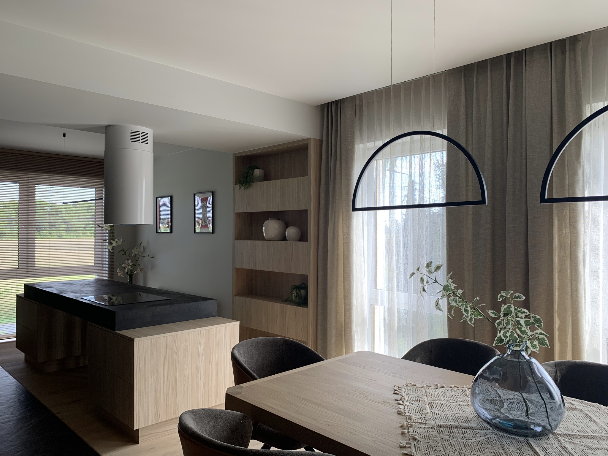 Nowoczesna aranżacja wnętrza: drewniana wyspa kuchenna z czarnym blatem, stół z dekoracyjnym bieżnikiem i wazonem, minimalistyczne lampy i zasłony w odcieniach szarości.