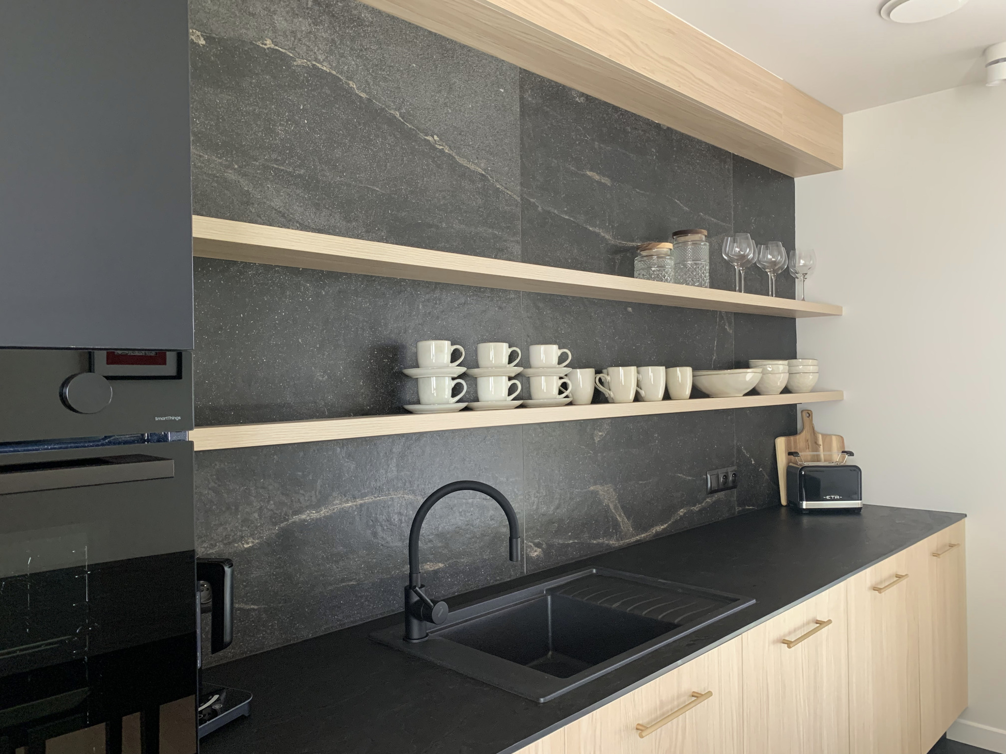 Nowoczesna kuchnia z czarnym blatem i armaturą, drewniane półki z zastawą, marmurowa ściana. Minimalistyczny design z akcentami naturalnego drewna i eleganckiej czerni.