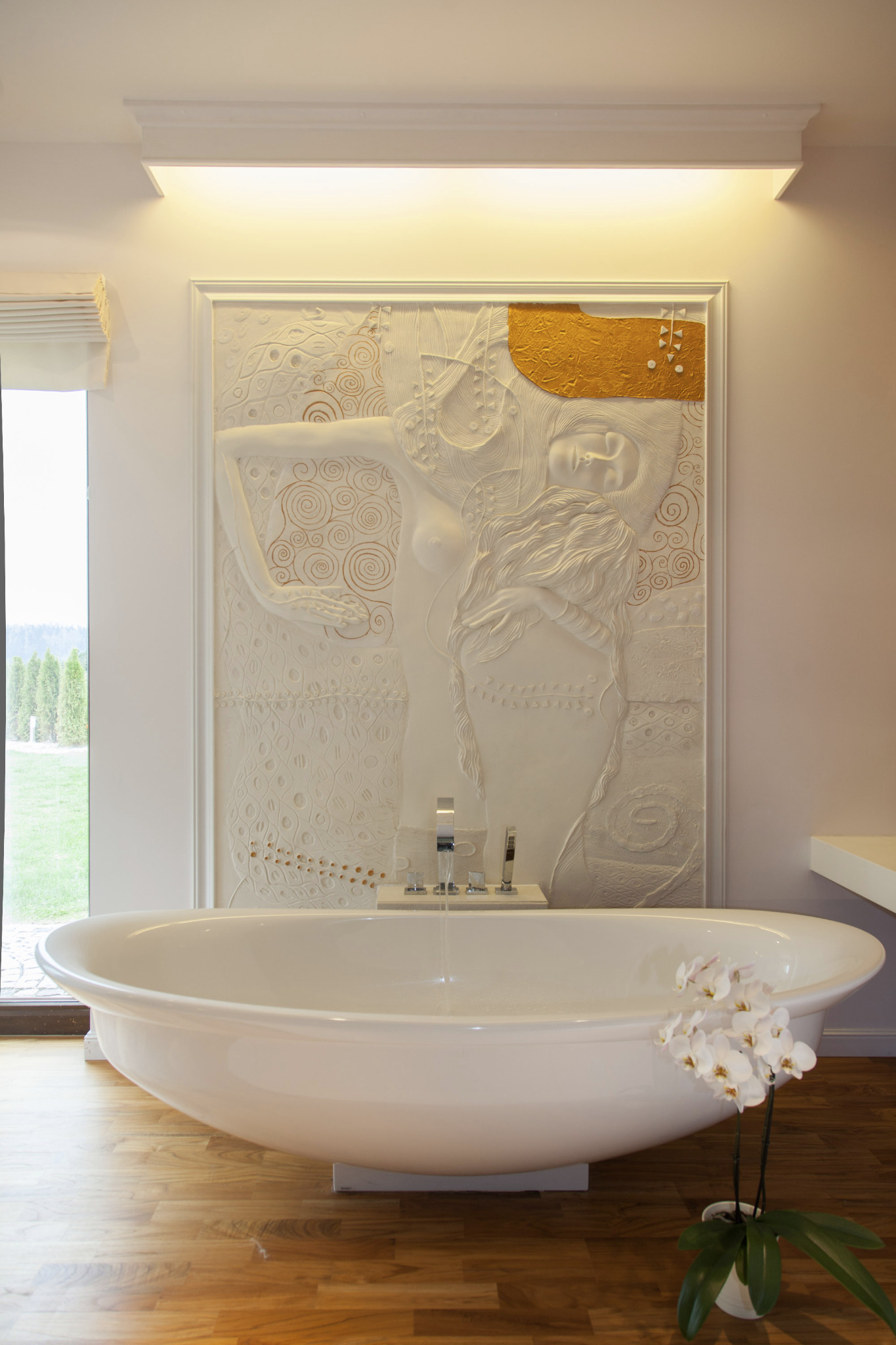 Biała, wolnostojąca wanna z kwiatami orchidei na drewnianej podłodze. W tle dekoracyjna płaskorzeźba nad wanną i okno z widokiem na zieleń.