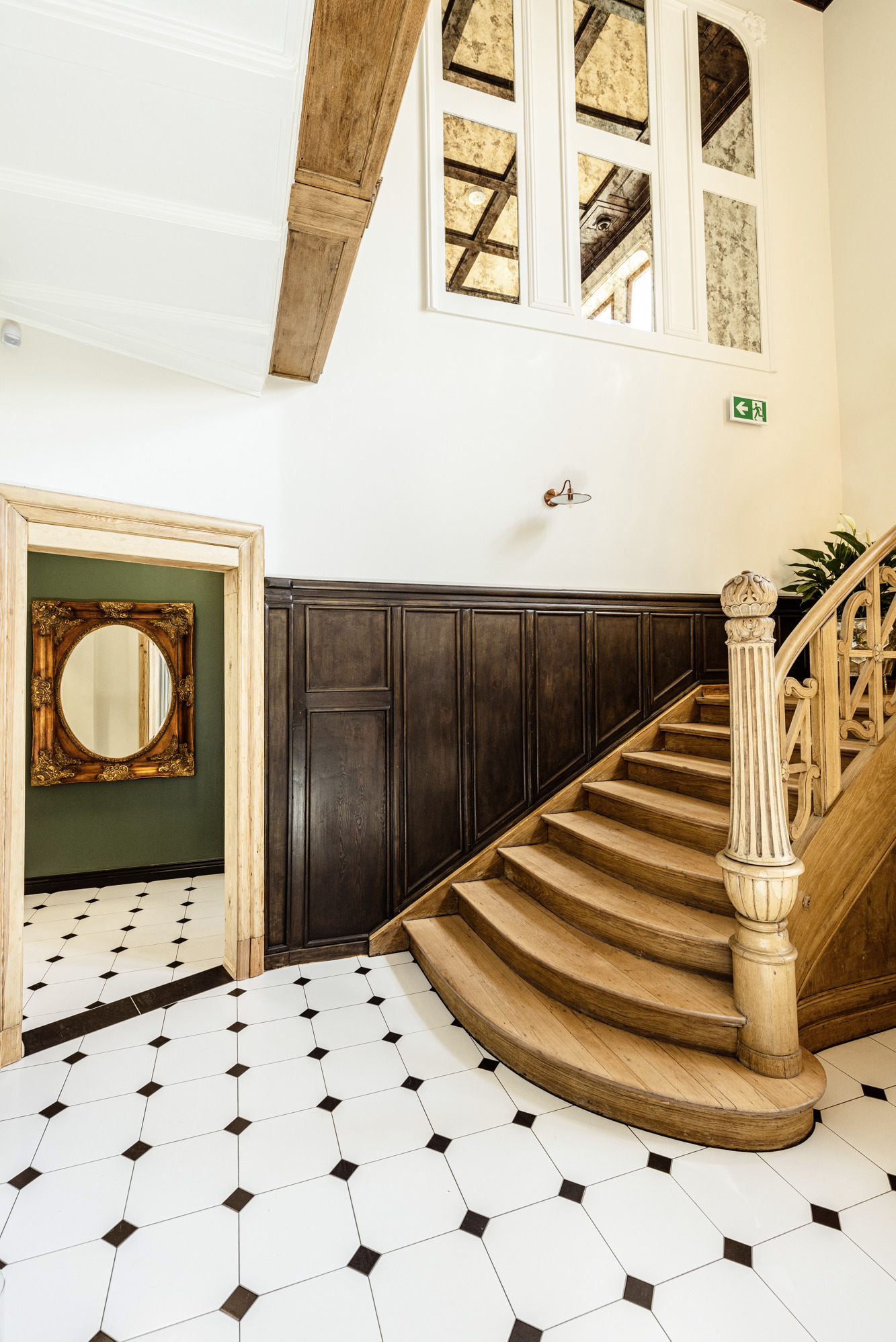 Stylowe wnętrze: drewniane schody z dekoracyjną balustradą, czarno-biała podłoga, ciemne panele ścienne i lustro w złotej ramie w otwartym przejściu.